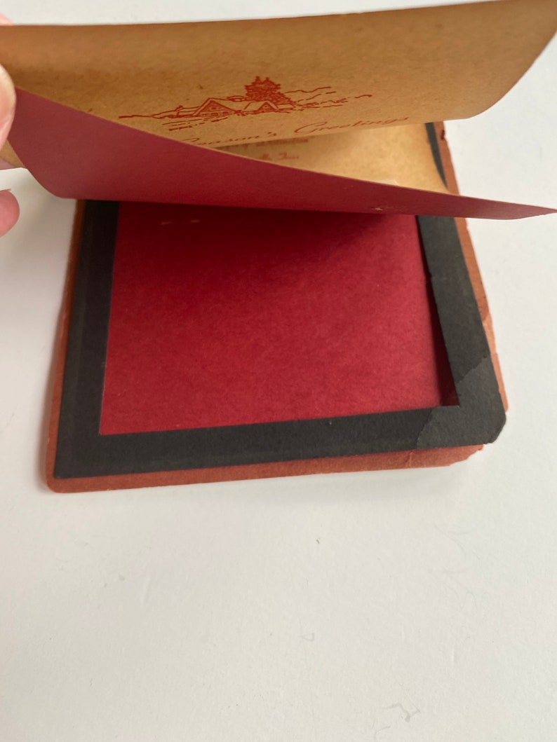 May include: A vintage greeting card with a red and brown color scheme. The card is open, revealing a red interior and a brown exterior with a red illustration and text. The card has a black border.