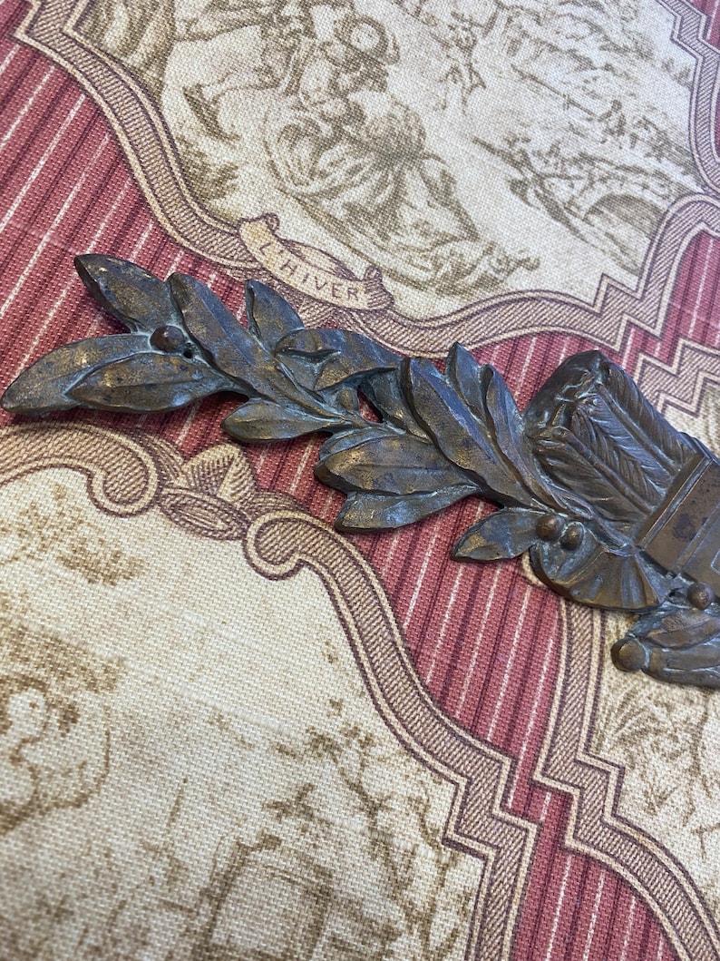 K&ouml;nnte beinhalten: Ein bronzenfarbener Metallobjekt mit einem stilisierten Zweig mit Bl&auml;ttern und Beeren. Das Objekt hat eine Schriftrolle mit der Aufschrift "L'HIVER".