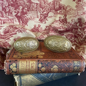 May include: Two antique gold-toned door knobs with ornate floral designs, resting on top of a vintage book with a red and gold cover.