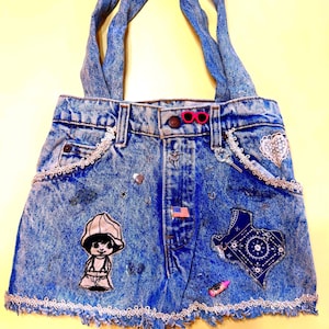 May include: A denim tote bag with a light blue wash, featuring a patchwork design with various embellishments. The bag has a cartoon character patch, a Texas-shaped patch, and decorative lace trim. The handles are made of the same denim material.
