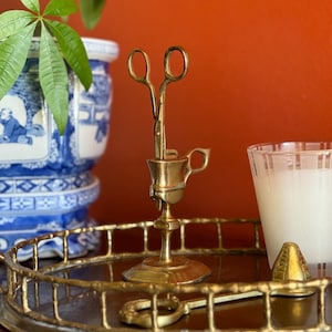 May include: A brass candle holder with a pair of scissors, a small spoon, and a pyramid-shaped object on a decorative tray. A white candle in a glass and a blue and white ceramic pot are also visible.