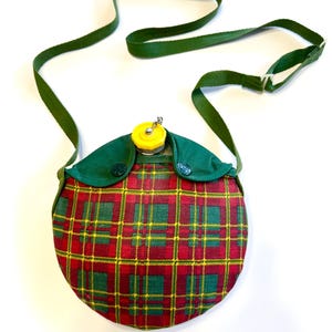 May include: A vintage canteen with a red, green, and yellow plaid pattern. The canteen has a green strap and a yellow cap. The canteen is round and has green fabric around the top. A classic camping accessory.