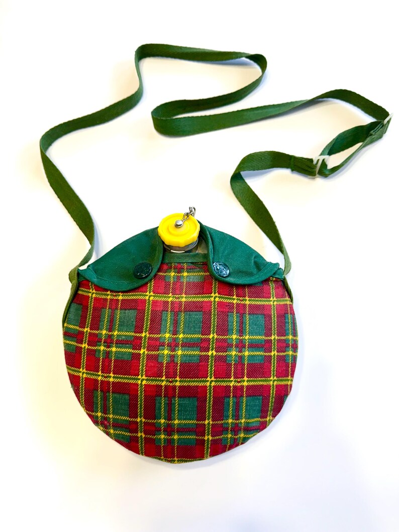 May include: A vintage canteen with a red, green, and yellow plaid pattern. The canteen has a green strap and a yellow cap. The canteen is round and has green fabric around the top. A classic camping accessory.