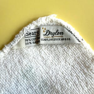 May include: Close-up of a white terry cloth towel with a tag that reads "Sears Drylon." The tag also indicates the towel is 70% cotton and 30% rayon. The Sears, Roebuck and Co. brand is also visible.