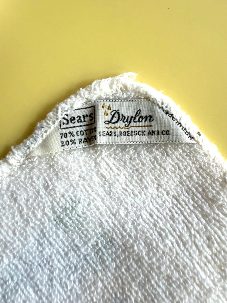 May include: Close-up of a white terry cloth towel with a tag that reads "Sears Drylon." The tag also indicates the towel is 70% cotton and 30% rayon. The Sears, Roebuck and Co. brand is also visible.