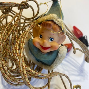 May include: A vintage Christmas light string with a smiling elf figurine holding a bell. The elf is wearing a blue and white hat and a blue and white outfit. The light string is tangled and has a black plug.