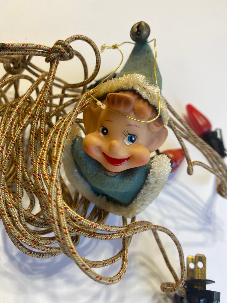 May include: A vintage Christmas light string with a smiling elf figurine holding a bell. The elf is wearing a blue and white hat and a blue and white outfit. The light string is tangled and has a black plug.