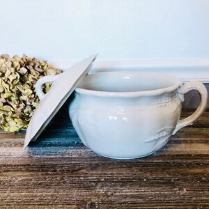 May include: White porcelain chamber pot with a lid. The pot has a decorative design of leaves and a scalloped rim. The lid has a single handle.