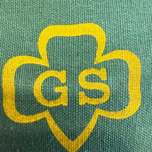 May include: Close-up of a green fabric with a yellow Girl Scout logo. The logo features a trefoil shape with the letters "GS" in the center. The fabric has a textured appearance.
