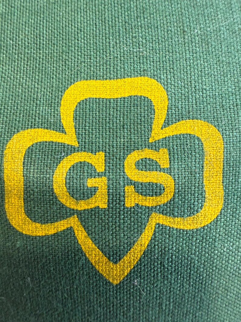 May include: Close-up of a green fabric with a yellow Girl Scout logo. The logo features a trefoil shape with the letters "GS" in the center. The fabric has a textured appearance.