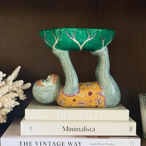 May include: A decorative ceramic bowl supported by a figure of a monkey. The bowl is green with pink accents, and the monkey figure is wearing a yellow garment with floral patterns. The figure is resting on a stack of books.