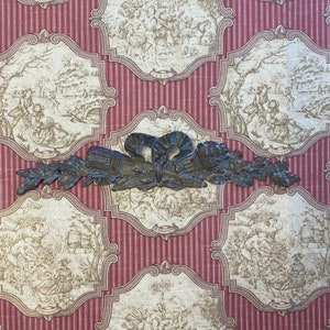 K&ouml;nnte beinhalten: Ein dekorativer Wandakzent aus Metall mit einem floralen Design und einer Schleife in der Mitte. Der Akzent ist in Bronze lackiert und hat eine rustikale Oberfl&auml;che.