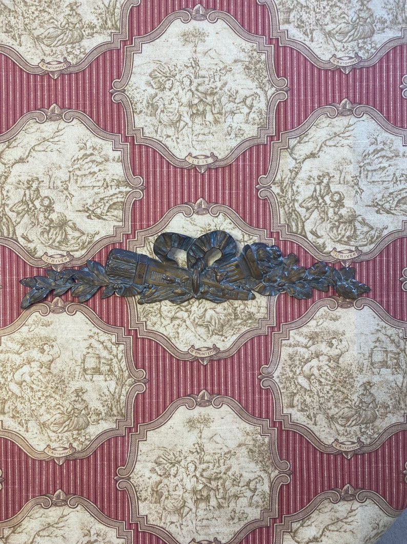 K&ouml;nnte beinhalten: Ein dekorativer Wandakzent aus Metall mit einem floralen Design und einer Schleife in der Mitte. Der Akzent ist in Bronze lackiert und hat eine rustikale Oberfl&auml;che.