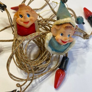 May include: A string of Christmas lights with elf faces on each bulb. The elves are wearing red and blue hats and have smiling faces. The lights are connected to a black plug.