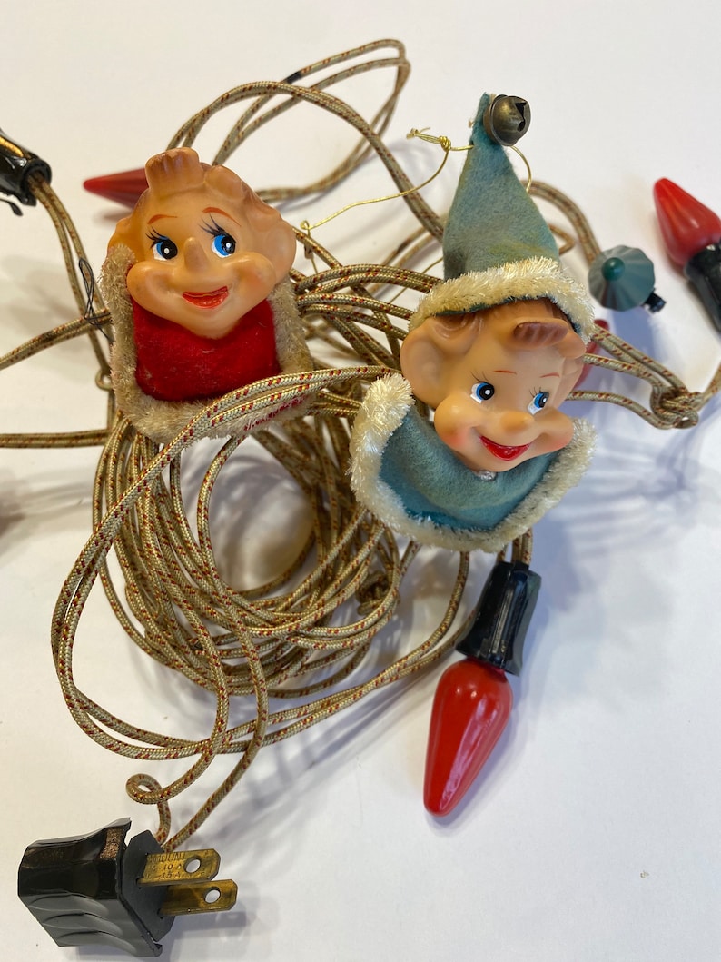May include: A string of Christmas lights with elf faces on each bulb. The elves are wearing red and blue hats and have smiling faces. The lights are connected to a black plug.