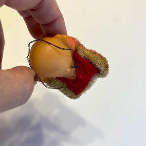 May include: A small, red and brown plush toy with a sewn-on face. The toy is in the shape of a person wearing a red and brown hat.