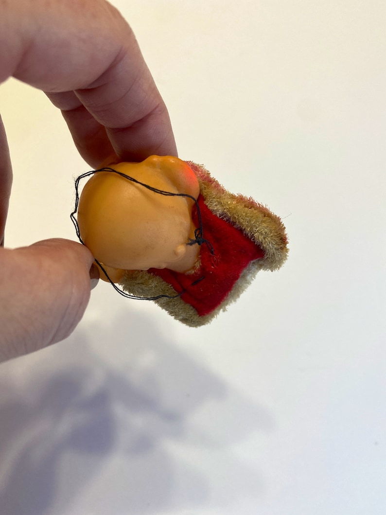 May include: A small, red and brown plush toy with a sewn-on face. The toy is in the shape of a person wearing a red and brown hat.