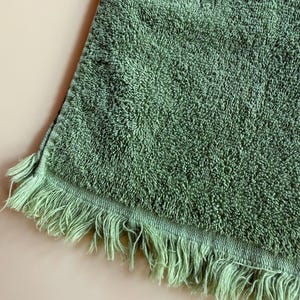 May include: A sage green bath towel with a textured surface and fringe detail along one edge. The towel is made of a soft, absorbent material, perfect for drying off after a shower or bath. The fringe adds a decorative touch.