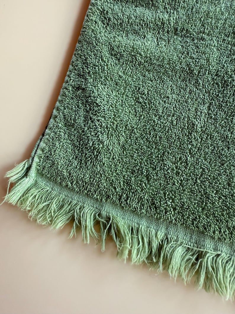 May include: A sage green bath towel with a textured surface and fringe detail along one edge. The towel is made of a soft, absorbent material, perfect for drying off after a shower or bath. The fringe adds a decorative touch.