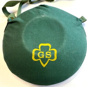 May include: A dark green canvas canteen cover with a matching adjustable shoulder strap. The cover features a yellow Girl Scout logo with the letters "GS" inside. The canteen cover is round and has a white plastic buckle.