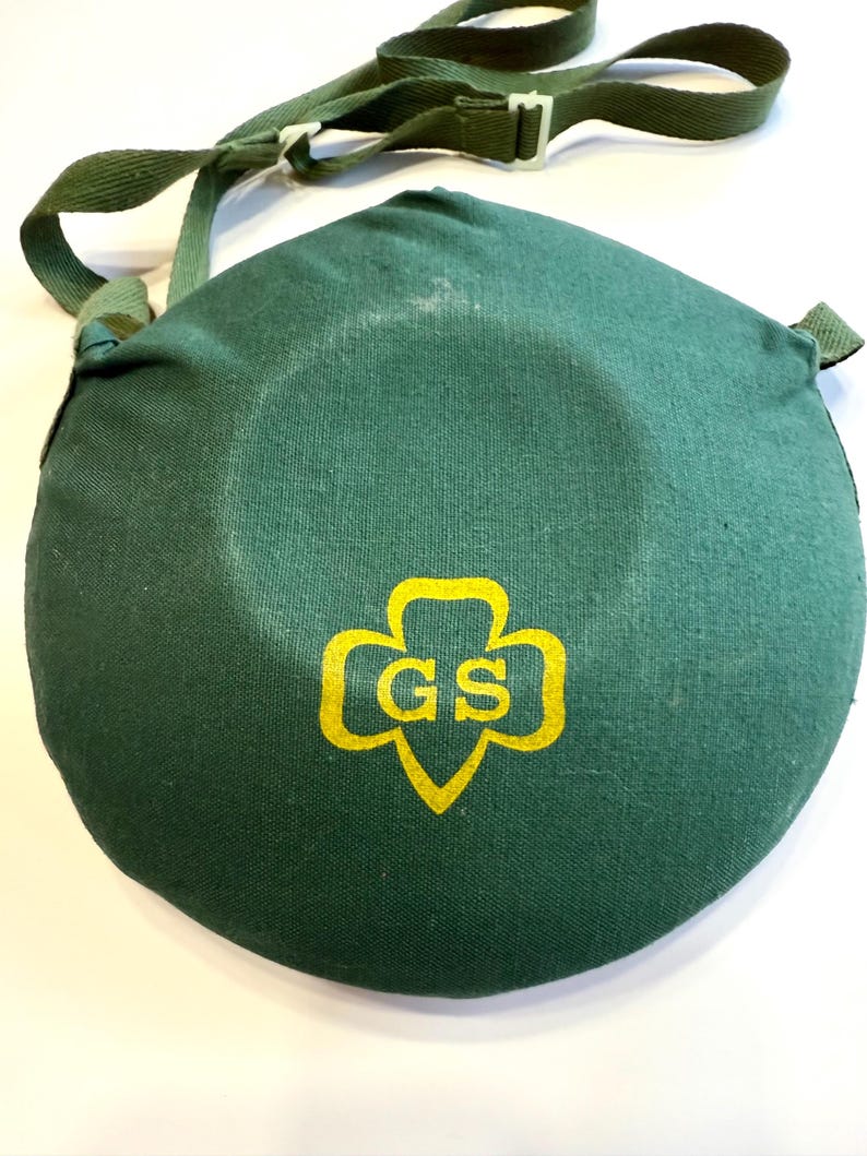 May include: A dark green canvas canteen cover with a matching adjustable shoulder strap. The cover features a yellow Girl Scout logo with the letters "GS" inside. The canteen cover is round and has a white plastic buckle.