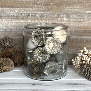 May include: A clear glass jar filled with vintage glass doorknobs of various shapes and sizes. The knobs are mostly clear with some having a slight amber tint. The jar sits on a white wooden surface.