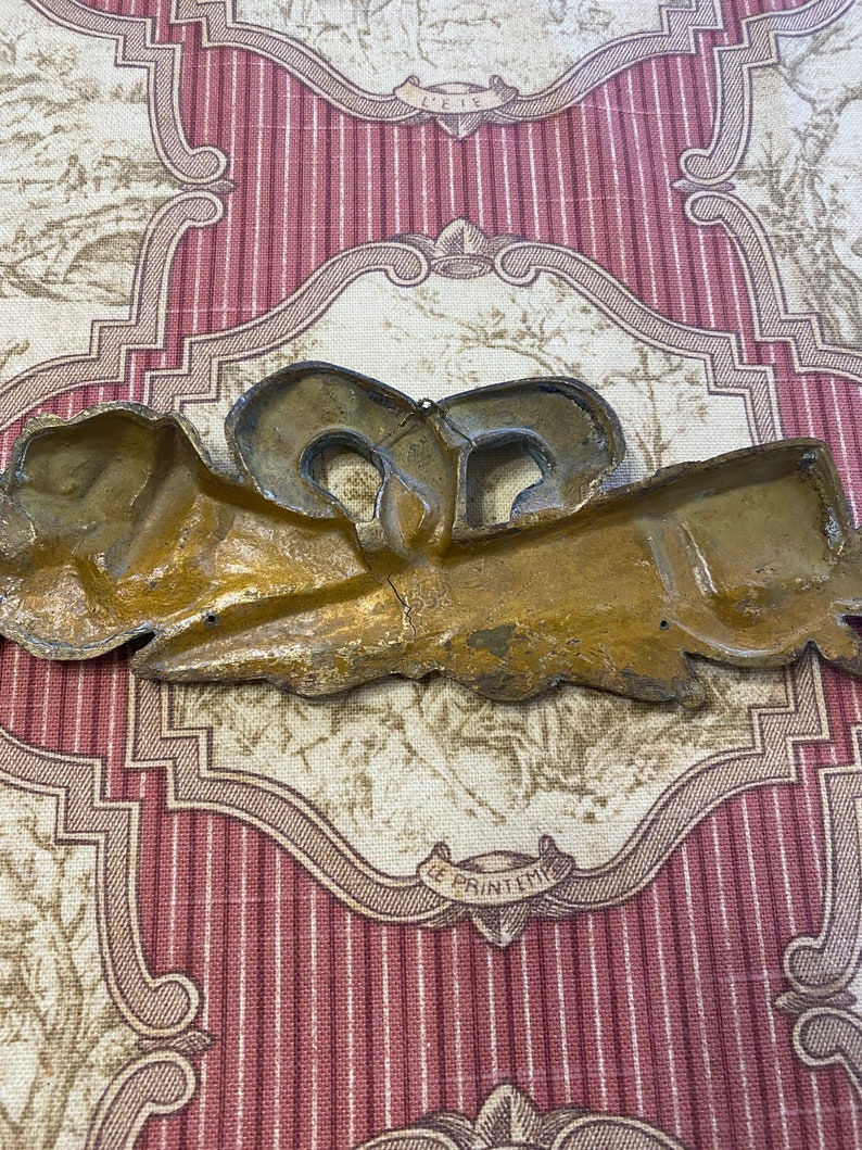 K&ouml;nnte beinhalten: Eine goldfarbene Wanddekoration aus Metall mit einem dekorativen Design. Das Design zeigt eine stilisierte Schleife mit zwei Schleifen und einem floralen Motiv. Die Dekoration hat eine patinierte Oberfl&auml;che und wirkt antik.