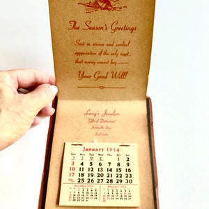 May include: An open, vintage booklet with a calendar for January 1954. The cover reads "The Season's Greetings" with a small illustration of a house. The booklet also features the text "Lang's Jewelers" and a calendar.