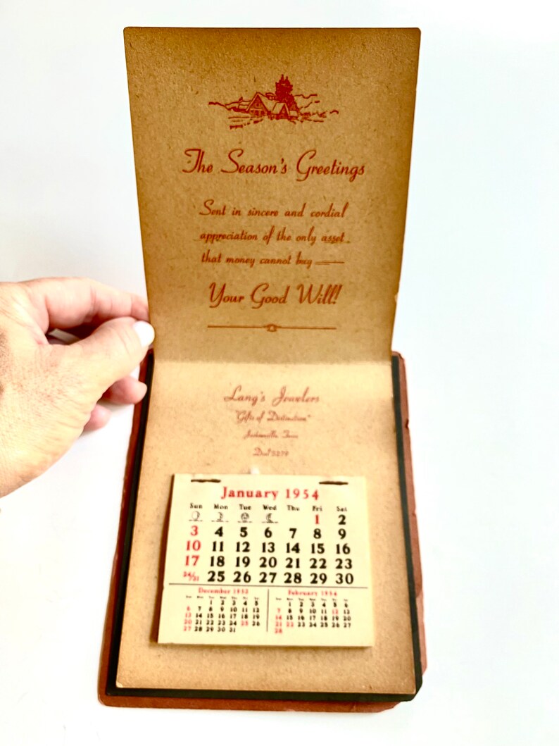 May include: An open, vintage booklet with a calendar for January 1954. The cover reads "The Season's Greetings" with a small illustration of a house. The booklet also features the text "Lang's Jewelers" and a calendar.