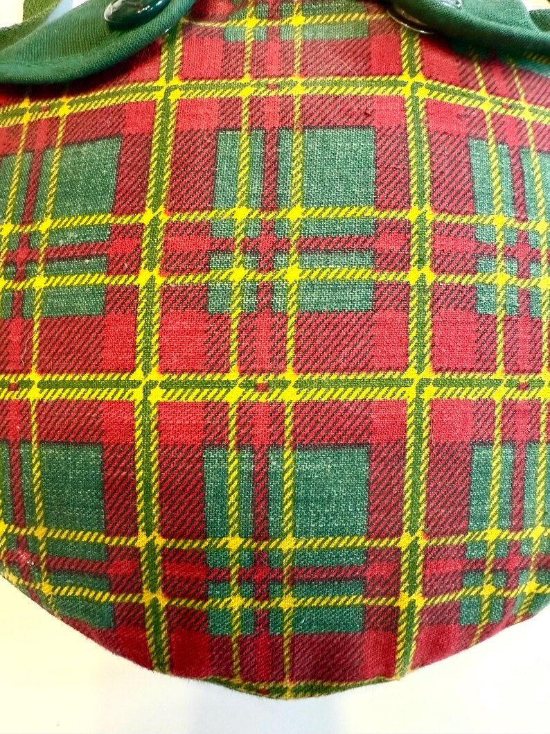 May include: Close-up of a plaid fabric with a pattern of red, green, and yellow lines. The fabric is a mix of red and green squares with yellow lines. A green fabric trim with two buttons is at the top.