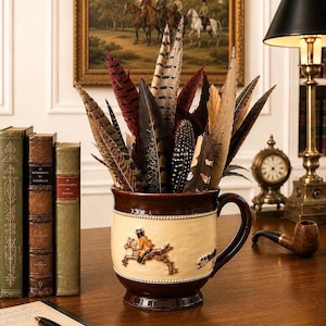 May include: A ceramic mug with a dark brown rim and base, featuring a cream-colored center band with a raised relief of a horse and rider. The mug is filled with an assortment of long, pointed feathers in shades of brown, tan, and deep red.