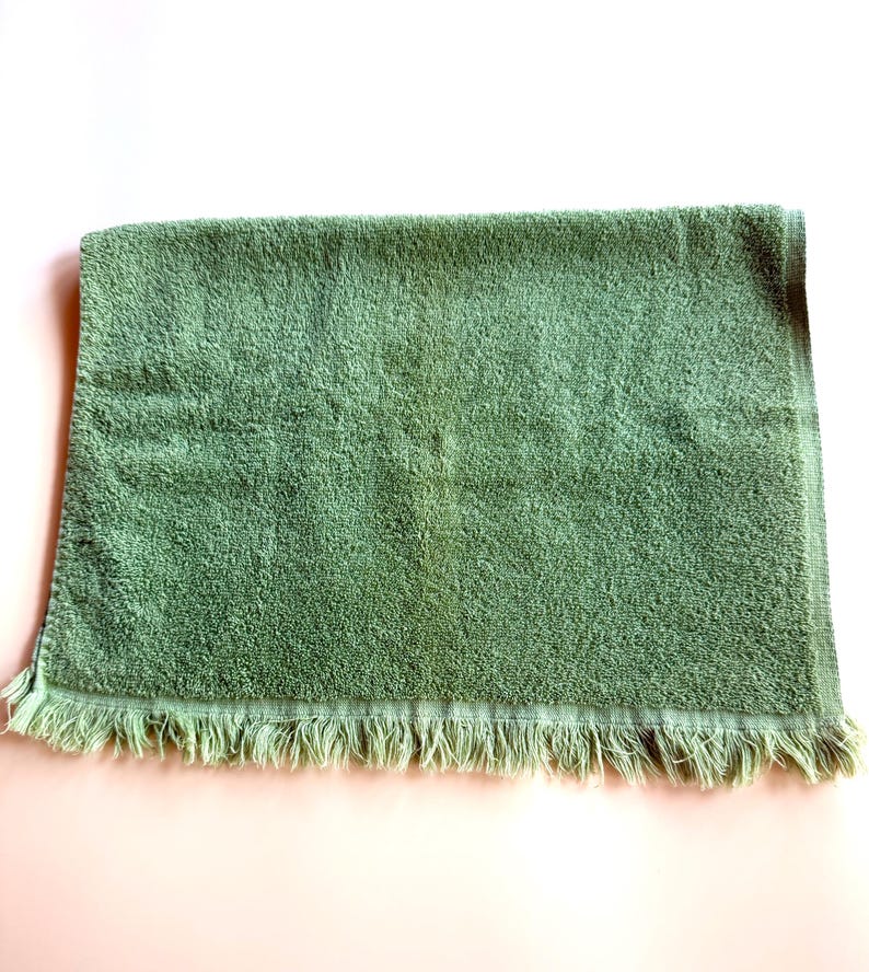 May include: A sage green bath towel with a fringed edge. The towel is rectangular and has a plush, absorbent texture. The fringe is a lighter shade of green. The towel is folded and lies on a light pink surface.