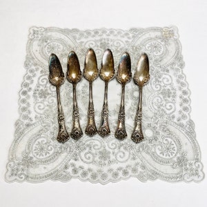 May include: Set of six silver-plated spoons with ornate handles. The spoons have a teardrop-shaped bowl and are arranged in a row on a white lace doily.