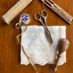 May include: A collection of sewing tools and supplies including a spool of thread, a wooden bobbin, a pair of scissors, a wooden needle threader, a measuring tape, and a piece of lace fabric.