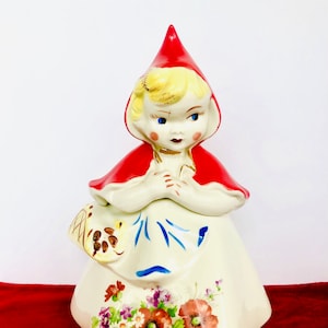 May include: A vintage ceramic cookie jar shaped like Little Red Riding Hood. The jar is painted with a white dress, red hooded cloak, and floral details. The jar has a blue and white pattern on the skirt and a basket of flowers in her hand.