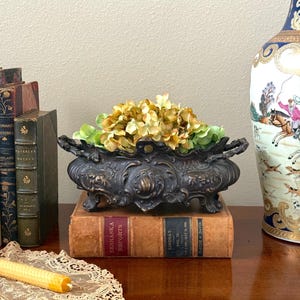 May include: An antique bronze-toned decorative planter filled with yellow and green flowers, resting on two vintage books. The planter has ornate detailing and handles. A tall, decorated vase is in the background.