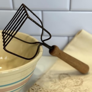 May include: A vintage potato masher with a wooden handle and a metal head. The masher is resting near a ceramic bowl with blue and pink stripes and a cream-colored cloth.