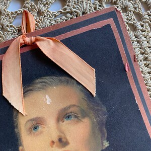 May include: Vintage portrait of a woman with blue eyes and red lipstick. The portrait is on a dark background with a peach-colored ribbon tied at the top. The edges of the portrait are trimmed in a dark brown color.
