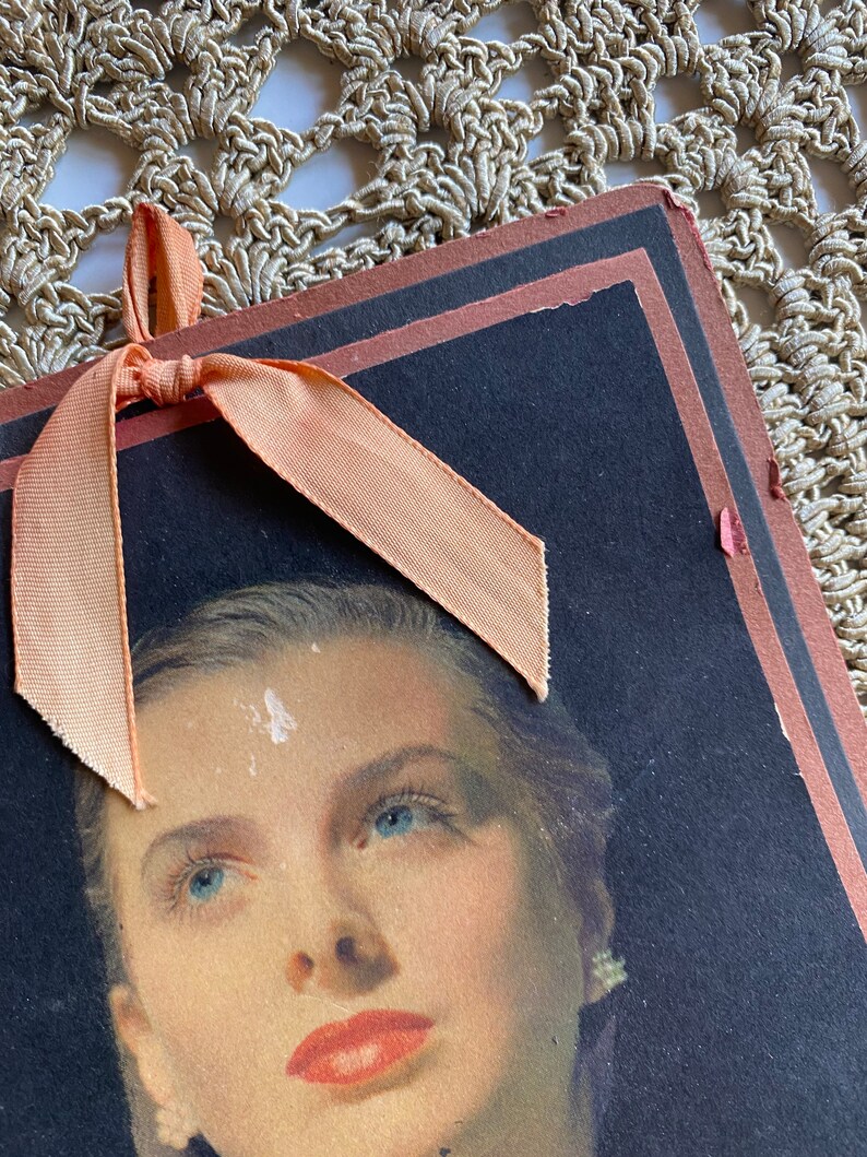 May include: Vintage portrait of a woman with blue eyes and red lipstick. The portrait is on a dark background with a peach-colored ribbon tied at the top. The edges of the portrait are trimmed in a dark brown color.