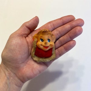 May include: A small, vintage, plastic figurine of a smiling elf with blue eyes. The elf is wearing a red and white hat and a brown fur collar.