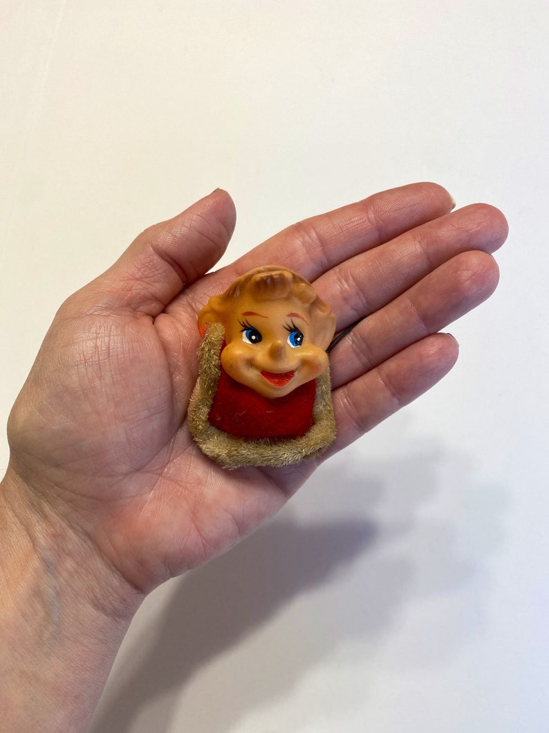 May include: A small, vintage, plastic figurine of a smiling elf with blue eyes. The elf is wearing a red and white hat and a brown fur collar.