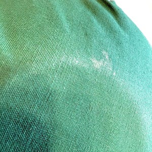 May include: Close-up of a textured, forest green fabric. The fabric has a woven pattern and appears to be a durable material. There are some white marks on the fabric. The image shows a portion of a larger item, possibly a bag or cushion.
