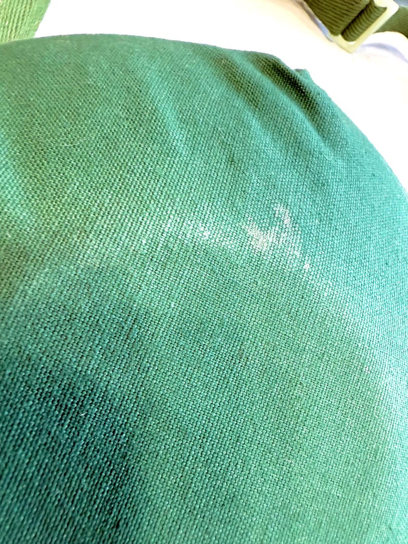 May include: Close-up of a textured, forest green fabric. The fabric has a woven pattern and appears to be a durable material. There are some white marks on the fabric. The image shows a portion of a larger item, possibly a bag or cushion.
