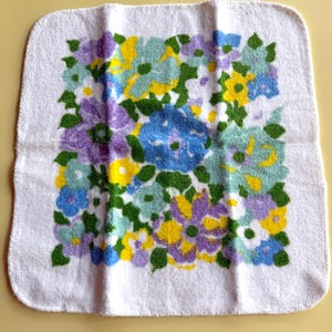 May include: White square towel with a floral pattern in shades of blue, yellow, purple, and green. The flowers are clustered together, creating a vibrant and colorful design. The towel is made of a soft, absorbent material, perfect for use in a bathroom or kitchen.