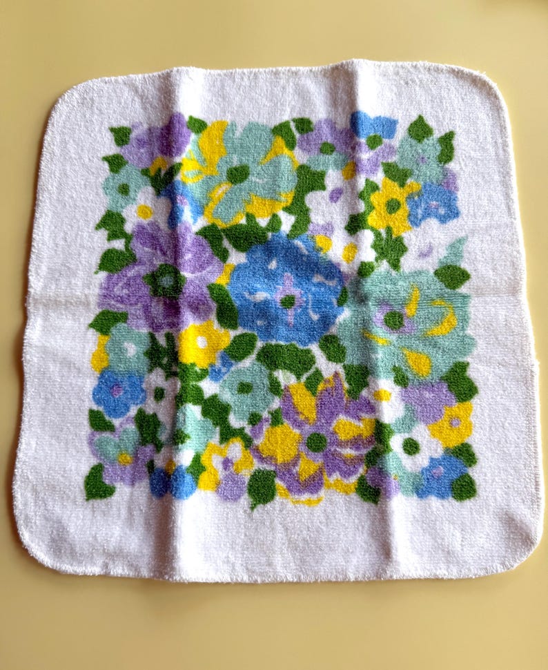 May include: White square towel with a floral pattern in shades of blue, yellow, purple, and green. The flowers are clustered together, creating a vibrant and colorful design. The towel is made of a soft, absorbent material, perfect for use in a bathroom or kitchen.