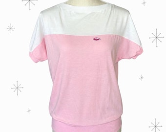 May include: A short-sleeved shirt featuring a color-blocked design with a white upper section and a pink lower section. The garment has a ribbed crew neckline and a ribbed hemline. A small embroidered pink crocodile logo is on the chest.