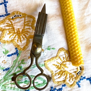 May include: Antique brass candle scissors with a sharp blade and two loops. The scissors are on a white embroidered tablecloth with a yellow honeycomb candle.