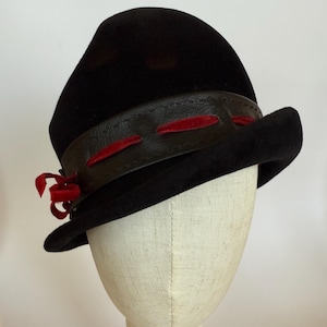 May include: A black cloche hat with a rounded crown and a wide brim. The hat features a dark brown leather band with red velvet accents and a small red ribbon bow on the side. The hat is displayed on a mannequin head.