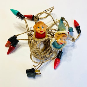 May include: A string of Christmas lights with elf faces on each bulb. The lights are red, green, and blue. The cord is brown with a red and white pattern.