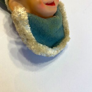 May include: A vintage elf ornament with a green felt hat and a white fur trim. The elf has a large nose, blue eyes, and a rosy-cheeked smile.